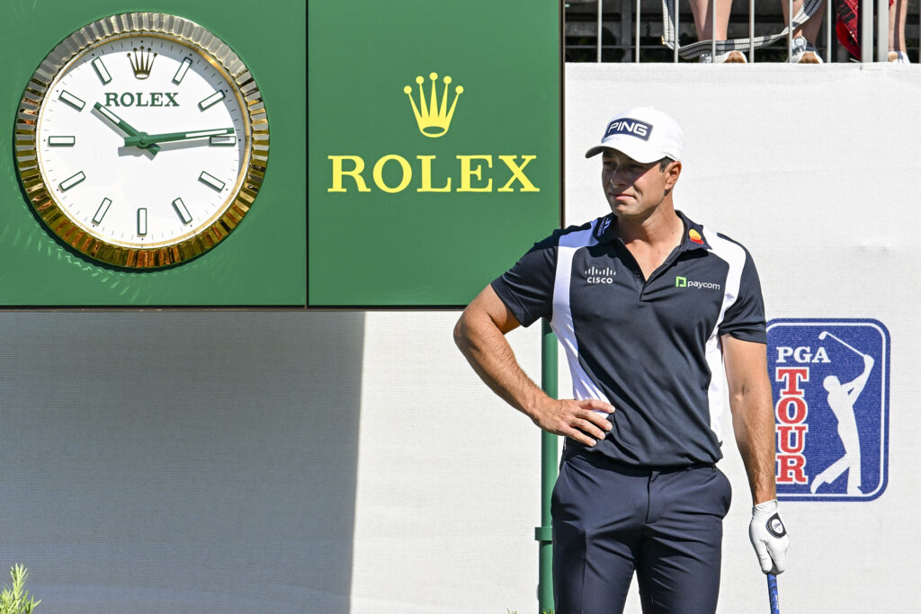 golfspelaren viktor hovland från norge står bredvid en grön rolexklocka på en golfbana iförd vit golfkeps och blåvit-randig piké