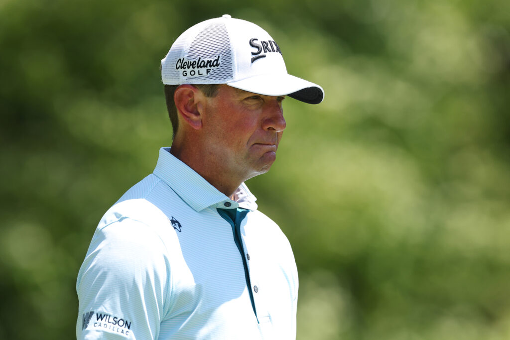 golfspelaren lucas glover i vit golfkeps och vit golfpiké på en amerikansk golfbana med lövträd