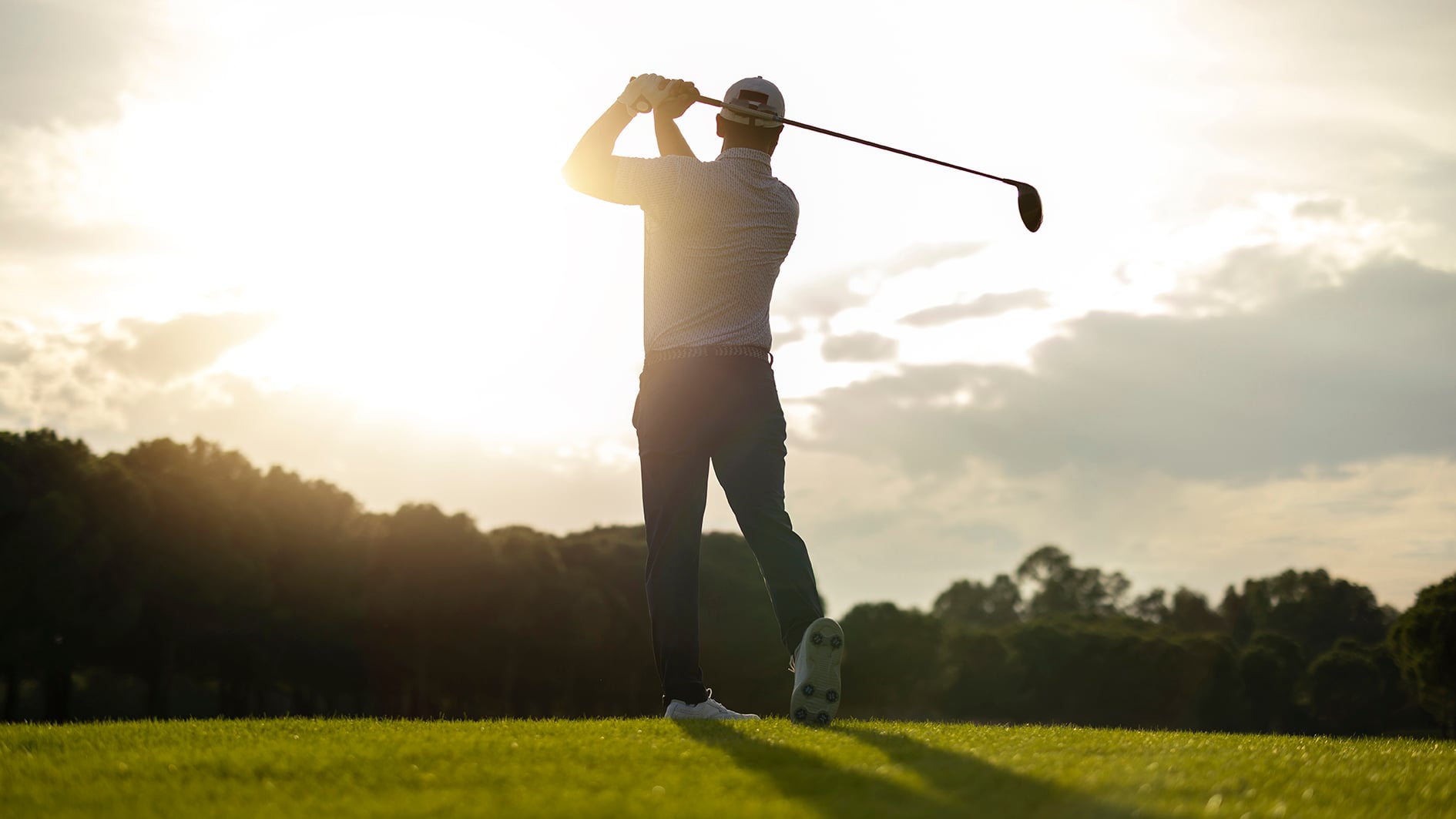 Silhouette of a golfer preparing to swing at sunrise on a bright fairway. Warm backlight and a calm outdoor atmosphere make the image suitable for sports, golf lifestyle, motivation, and leisure themes.