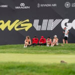 golfspelare sitter i gräset på en golfbana nedanför en läktare.