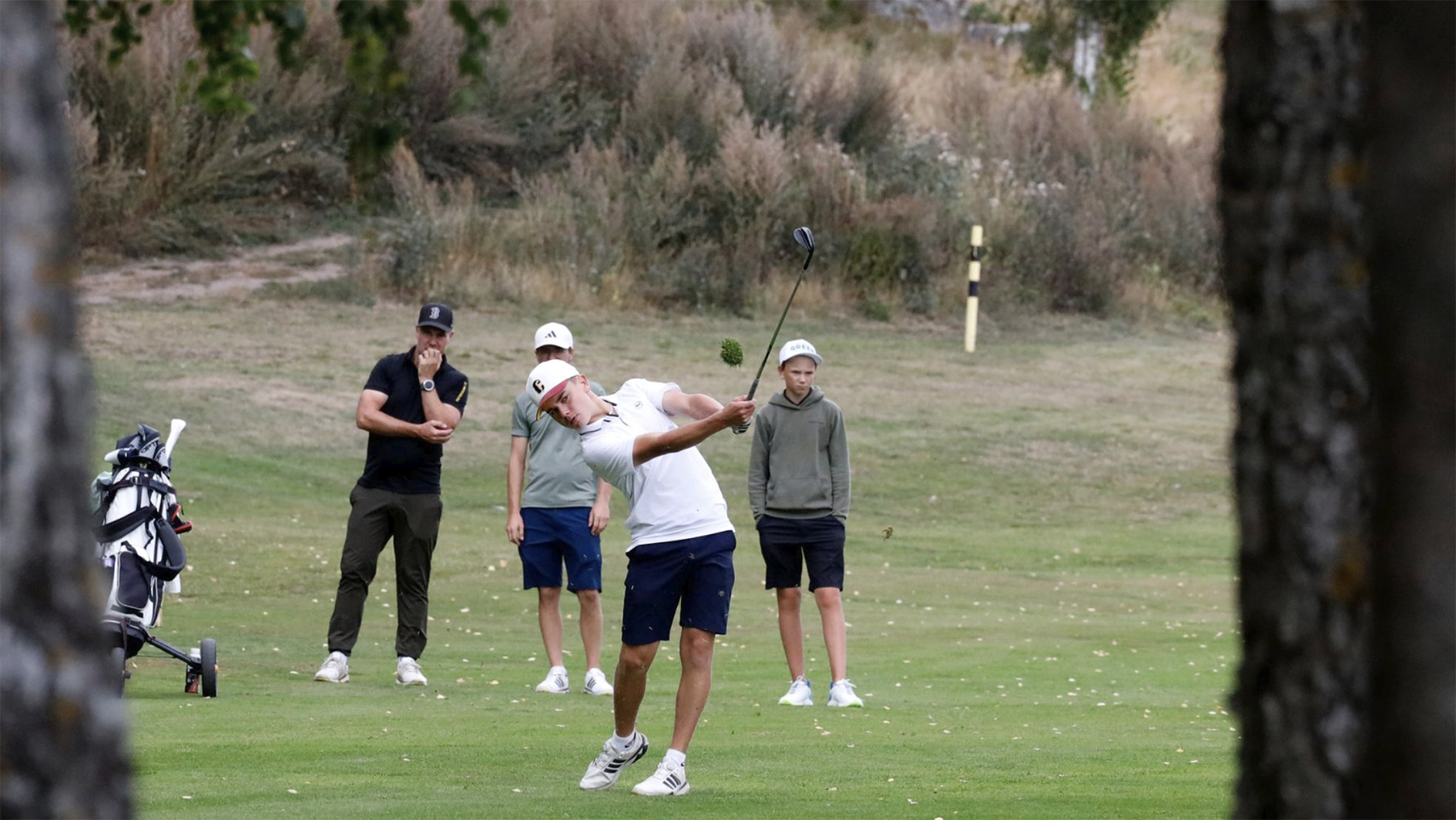 En pojke slår en golfboll med en driver medan tre andra personer tittar på, på en golfbana med gräs och buskar i bakgrunden.